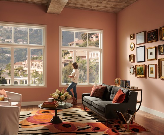 A woman standing against windows looking out to a picturesque mountain view. The room is peach colored with dramatic lighting. The room is a living room with a couch and chairs for a sitting area.